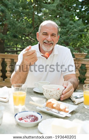 Portrait of a senior man sitting at a table having breakfast in a luxury hotel garden on holiday. Mature people eating and drinking healthy food and relaxing, smiling. Outdoors lifestyle.