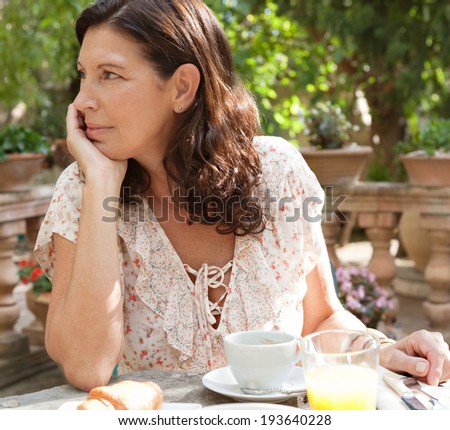 Attractive mature woman sitting at a table in a luxury villa hotel garden having a healthy continental breakfast during a sunny day on holiday. Senior living and food eating, outdoors lifestyle.