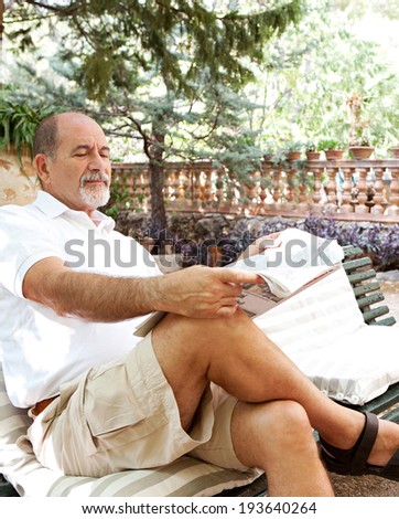 Senior man relaxing in a luxury holiday villa green garden, sitting on a bench and reading a newspaper while on vacation in a Mediterranean countryside hotel. Mature travel and outdoors lifestyle.