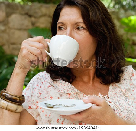 Portrait of a senior woman in a luxury hotel garden during a sunny day on holiday drinking coffee and relaxing on vacation. Mature people drinking hot beverages, outdoors. Senior people lifestyle.