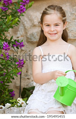 Close up portrait of a beautiful girl child sitting in a holiday home garden holding a small watering can and smiling at the camera during  a sunny day. Childhood summer vacation lifestyle, outdoors.