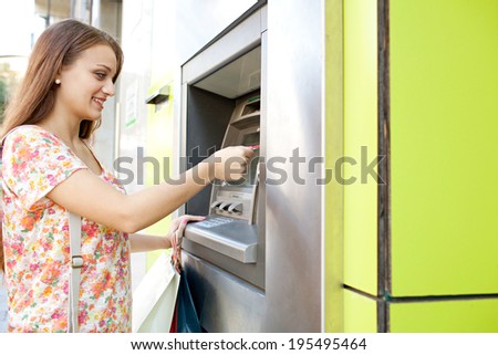 Side portrait of an attractive young woman using a cash point machine with her credit card to withdraw money while shopping in a city and carrying paper bags. Consumerism and lifestyle, outdoors.