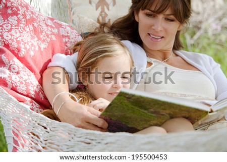 Close up portrait of a beautiful young mother and her young daughter laying down in a hammock relaxing together, reading a children stories book during a sunny summer day. Family lifestyle outdoors.