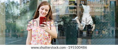 Panoramic view of an attractive joyful woman shopping in a city by a fashion store window with reflections, using a smartphone to take a selfie photo, outdoors. Consumerism and technology lifestyle.