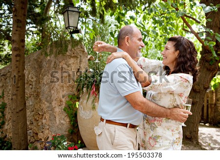 Mature couple dancing while drinking champagne in a luxury hotel garden on a summer holiday and dancing together holding their glasses. Senior people enjoying life and retirement, outdoors lifestyle.