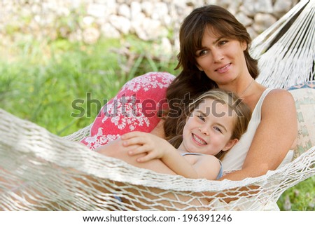 Joyful mother and young daughter relaxing together on a hammock in a holiday home garden with green grass during a sunny summer day. Family activities and lifestyle outdoors.