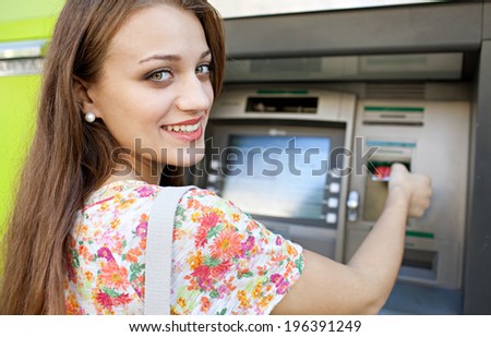Close up rear view of an attractive young woman using a cash point machine to withdraw money, inserting her credit card, turning and smiling during a sunny day outdoors. Finance and lifestyle.