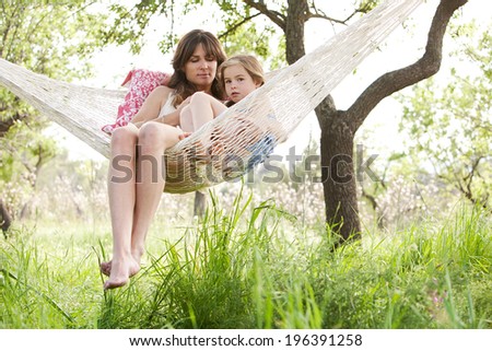 Wide view of a mother and daughter relaxing together being thoughtful sitting in a hammock, hugging and lounging during a sunny summer day in a holiday home garden with grass and trees, lifestyle.