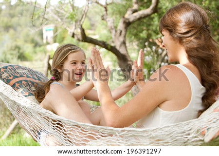 Young daughter and mother sitting together on a hammock in a holiday home garden playing games and clapping their hands, having fun during a sunny summer vacation. Active family lifestyle.