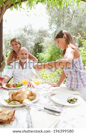Joyful family sitting at a healthy lunch table in a holiday villa green garden, relaxing during a summer day eating fresh food and enjoying life with child pouring juice. Outdoors eating lifestyle.