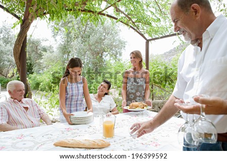 Joyful family members setting a food table outdoors together, placing plates and glasses during a sunny summer holiday day in a vacation villa home garden, outdoors. Teamwork and eating lifestyle.