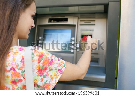 Close up rear view of an attractive young woman using a cash point machine to withdraw money, inserting her credit card and smiling during a sunny day outdoors. Finance and lifestyle.