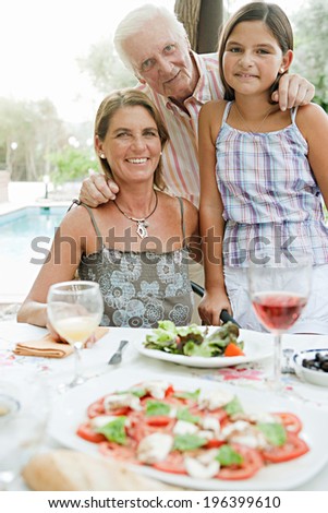 Portrait of a happy grandfather with his young granddaughter and daughter smiling during a sunny summer day on a holiday home green garden while having lunch outdoors. Family on vacation, lifestyle.