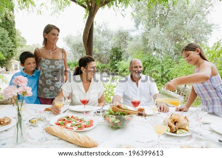 Joyful family sitting at a healthy lunch table in a holiday villa green garden, relaxing during a summer day eating fresh food and enjoying life with girl pouring juice. Outdoors eating lifestyle.