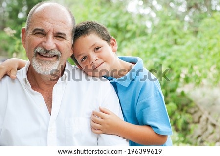 Close up portrait of a young son and his grandfather relaxing and hugging in a green garden during a sunny day, enjoying each others company and smiling joyfully. Outdoors active lifestyle.