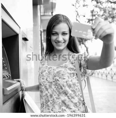 Black and white portrait of attractive woman using a cash machine point to withdraw money, and showing off her credit card joyfully, carrying shopping bags during a sunny day in the city, lifestyle.