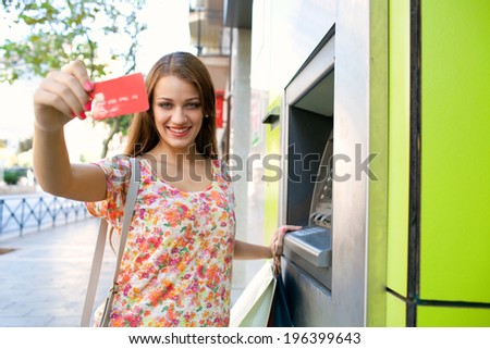 Portrait of an attractive young woman using a cash machine point to withdraw money, and showing off her credit card joyfully while carrying shopping bags during a sunny day in the city, lifestyle.