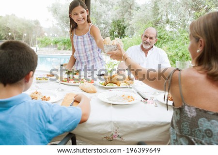 Joyful family sitting at a healthy lunch table in a holiday villa green garden, relaxing during a summer day eating fresh food and enjoying life with girl pouring juice. Outdoors eating lifestyle.