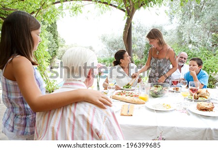 Joyful family group sitting at a healthy lunch table in a holiday villa green garden on vacation, relaxing during a summer day eating fresh food and enjoying life. Outdoors eating lifestyle.
