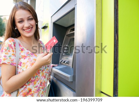 Joyful portrait view of an attractive young woman using a cash machine to withdraw cash money with her credit card, having fun and smiling in a city street. Outdoors finance and lifestyle.
