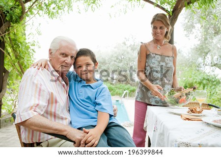 Grandson and grandfather sitting together at a healthy lunch table outdoors in a holiday villa green garden, smiling and relaxing by the swimming pool and eating healthy salad. Vacation lifestyle.