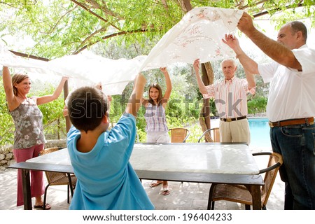 Large family group setting up a lunch table together throwing a table cloth over in teamwork, getting ready for eating on a sunny day on holiday in a summer vacation home garden, outdoors lifestyle.