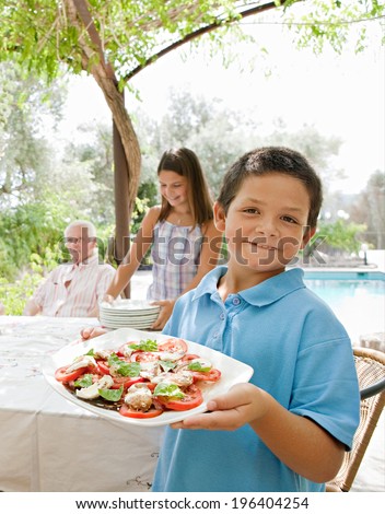 Joyful family preparing a table outdoors together, placing plates, glasses and healthy food during a sunny summer holiday day in a vacation villa home garden, outdoors. Teamwork and eating lifestyle.
