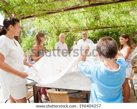 Large family group setting up a lunch table together throwing a table cloth over in teamwork, getting ready for eating during a sunny day on holiday in a home vacation garden, outdoors lifestyle.