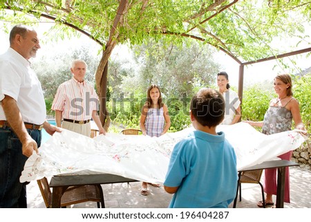 Large family group setting up a table cloth together in teamwork, getting ready for eating lunch outdoors during a sunny day on holiday in a summer villa vacation home garden, relaxing lifestyle.
