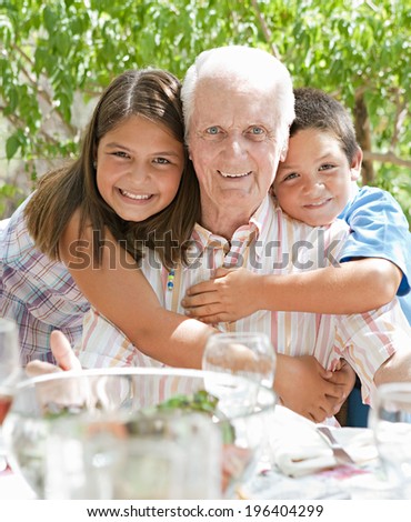 Portrait of a happy grandfather with his joyful grand children hugging him during a sunny summer day on a holiday home green garden while having lunch outdoors. Family relaxing on vacation, lifestyle.