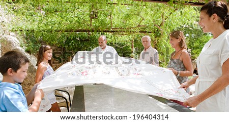 Large joyful family setting a food table together outdoors and coordinating placing a table cloth on during a sunny summer holiday day in a vacation home garden, outdoors. Teamwork and lifestyle.