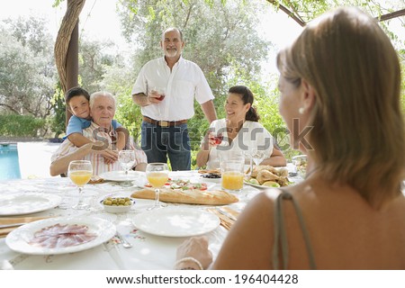 Group of family members enjoying a Spanish tapas meal in a villa green garden while on a holiday in a summer home with swimming pool, eating and drinking together during a summer day, outdoors.