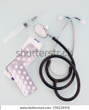 Still life close up detail view of a doctor stethoscope laying on a plain blue background with a syringe and prescription pills in a surgery interior. Medical and hospital equipment.