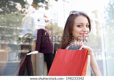Close up portrait of a beautiful young woman carrying shopping bags and walking by a fashion store in a city street, turning to look at the camera smiling. Consumer lifestyle outdoors.