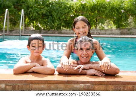 Family portrait of a father and his son and daughter enjoying the swimming pool together leaning on the stone edge and smiling at the camera during a summer holiday outdoors. Active family lifestyle.