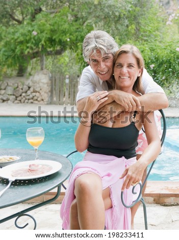 Husband and wife relaxing together and hugging while eating and drinking on a summer holiday in a vacation home, outdoors, hugging and smiling by a swimming pool. Active mature lifestyle.