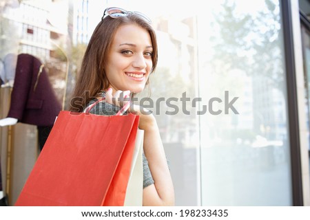 Close up portrait of a beautiful young woman carrying shopping bags and walking by a fashion store in a city street, turning at the camera smiling during a sunny day. Consumer lifestyle outdoors.