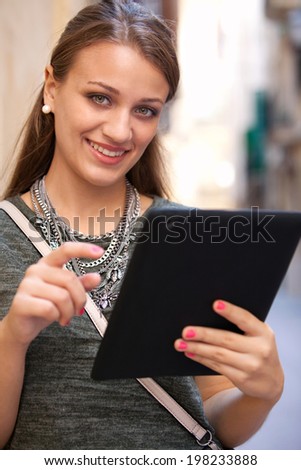 Close up portrait of an attractive young woman leaning on a wall using a digital tablet while visiting a characterful city street and smiling during a sunny day outdoors. Technology and lifestyle.