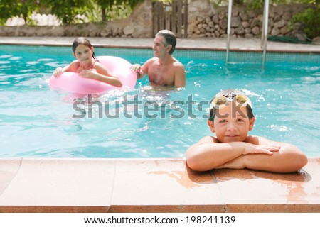 Father and children playing games having fun in a holiday swimming pool, with boy leaning on pool edge smiling and relaxing during a sunny summer day. Family activities and fun outdoors, lifestyle.