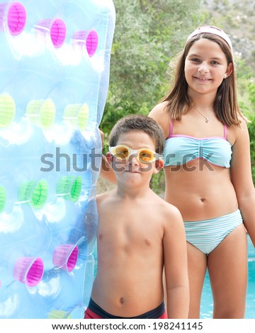 Colorful portrait of a young brother and sister standing together at edge of a swimming pool, smiling and holding a blue inflatable bed during a summer holiday in a hotel. Active vacation lifestyle.