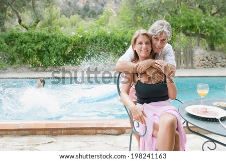 Father and mother relaxing together and hugging while on a summer holiday in a vacation home, outdoors, with children playing and having fun in the swimming pool splashing water. Active lifestyle.