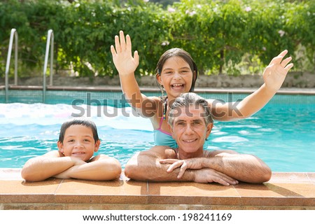 Family portrait of a father and his son and daughter enjoying the swimming pool together leaning on the stone edge and smiling at the camera during a summer holiday outdoors. Active family lifestyle.