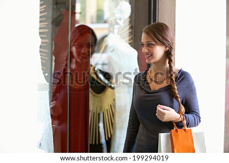 Portrait of a beautiful young woman visiting a luxury store display window with manikins and exclusive fashion during a sunny holiday, smiling. Shopping and consumer lifestyle, outdoors.
