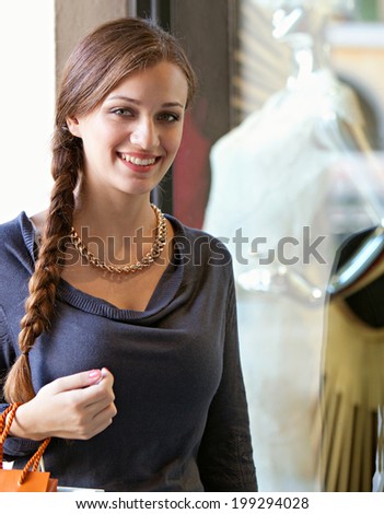 Portrait of a beautiful young woman visiting a luxury store display window with manikins and exclusive fashion during a sunny holiday, smiling. Shopping and consumer lifestyle, outdoors.