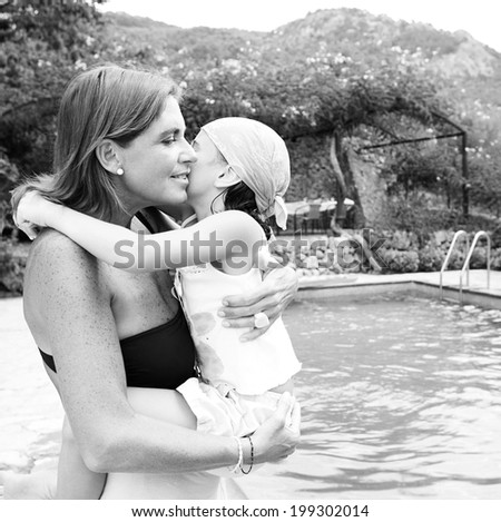 Black and white portrait of a mother carrying her daughter in her arms while visiting a summer holiday hotel villa, hugging and kissing being loving and close. Family vacation lifestyle, outdoors.