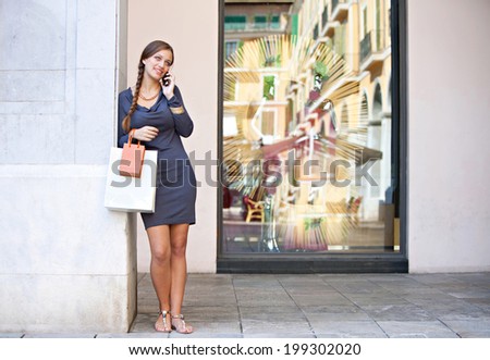 Stylish and beautiful young woman carrying shopping bags and leaning on a column using her smartphone cell to make a call while in a luxurious shopping mall, lifestyle. Consumerism and technology.