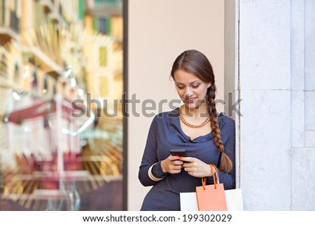 Stylish portrait of an elegant young woman carrying shopping bags and leaning on a column using her smartphone cell while visiting a luxurious shopping mall, lifestyle. Consumerism and technology.