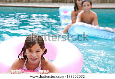 Close up portrait of a young girl enjoying a summer holiday in a swimming pool with her brother and sister, holding to the edge of the pool with an inflatable ring, smiling. Active vacation lifestyle.
