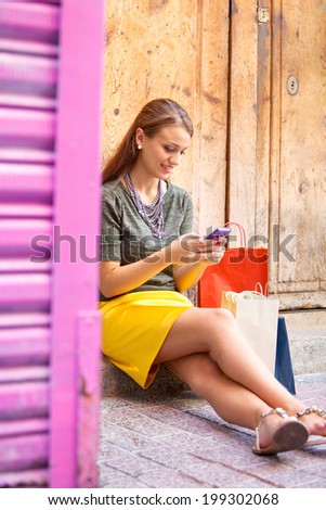 Close up portrait of an attractive young woman sitting by an old textured wooden door with shopping bags, smiling using a smartphone to network on vacation. Outdoors technology and lifestyle.