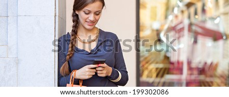 Panoramic portrait of a beautiful young woman carrying shopping bags and leaning on a column using her smartphone cell while visiting a luxurious shopping mall, lifestyle. Consumerism and technology.